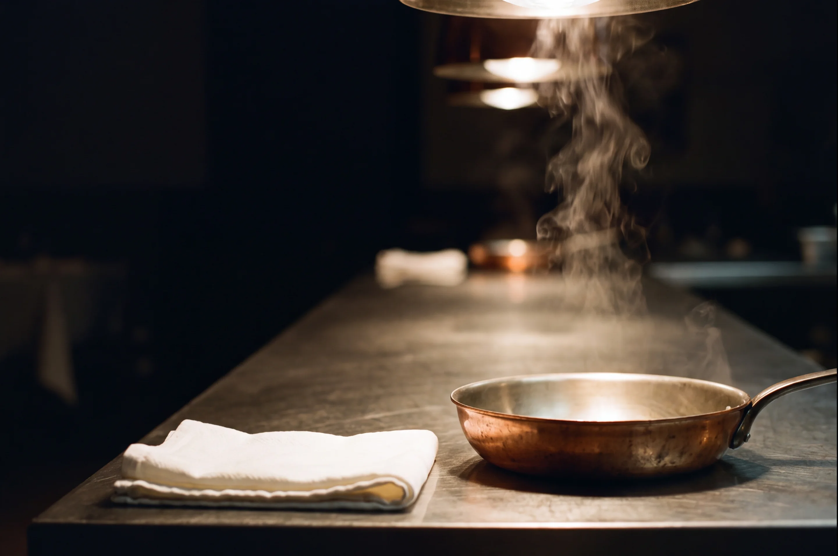 Une passe de cuisine professionnelle dans la dernière minute calme avant le service : plan de travail en acier brossé, lampe chauffante, colonne de vapeur, linge plié, poêlon de cuivre. Image générée du métier — aucun établissement identifiable, aucune campagne, aucun travail livré.